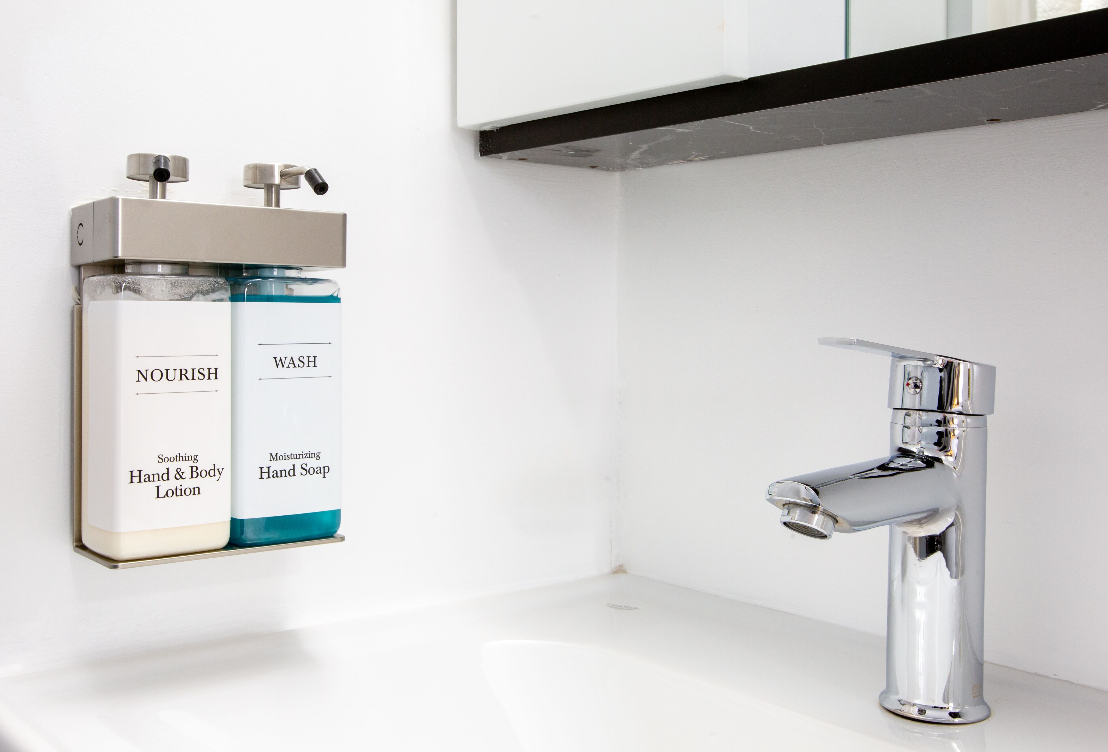 Une salle de bain moderne avec un robinet chromé et deux distributeurs de savon liquide portant les inscriptions « Nourrir » et « Laver ».