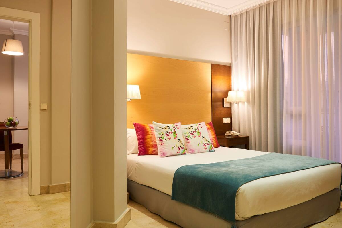 Habitación de hotel con cama, lámparas de noche, mesita de noche y vistas a la ciudad a través de cortinas translúcidas.