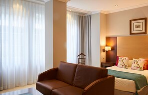 Habitación de hotel con sofá de cuero marrón, cama con almohadas estampadas, mesita de noche con lámpara y cortinas translúcidas.