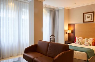 Ein Hotelzimmer mit einer braunen Ledersofa, einem Bett mit blumigen Kissen, einem Nachttisch mit Lampe und durchsichtigen Vorhängen.