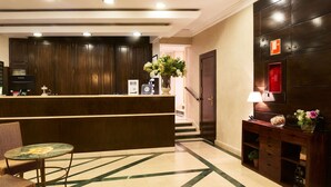 Zona de recepción de hotel con un mostrador de madera oscura, una mesa de vidrio con sillas, un jarrón con flores y una estantería con adornos.
