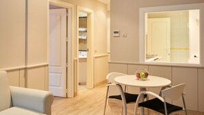Una habitación de hotel moderna con zona de comedor que incluye una mesa redonda y dos sillas, un frutero pequeño sobre la mesa, y vista al baño por una ventana.