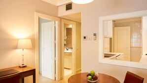 Habitación de hotel con zona de comedor que incluye una mesa redonda con frutero, una silla roja, una lámpara y vista a la cocina por una ventana.