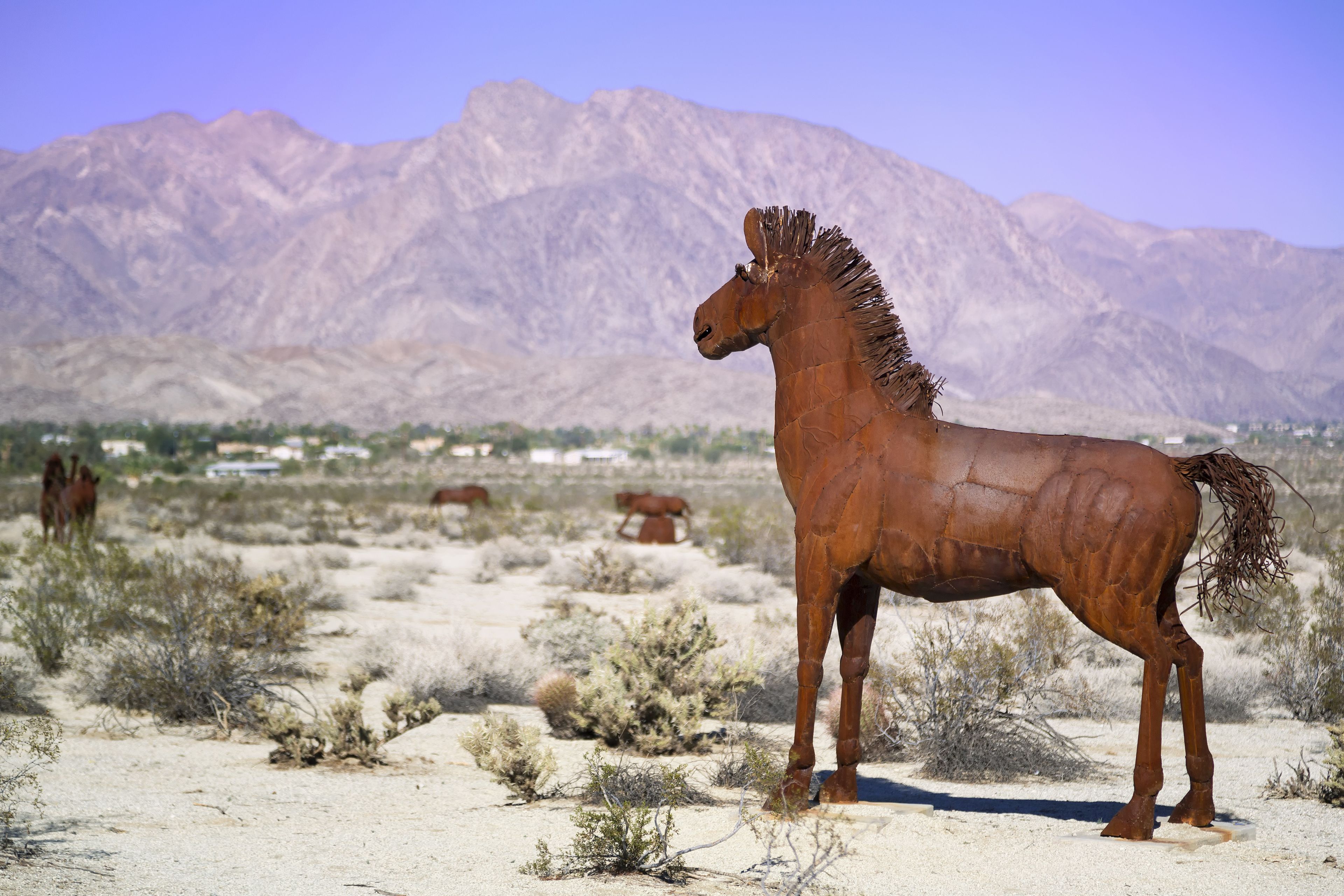 Una gran estatua de un caballo color óxido en un paisaje desértico con montañas al fondo.