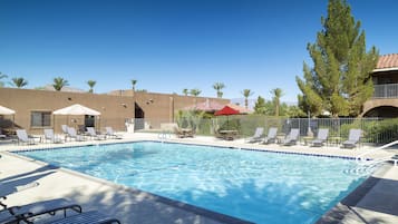 Ein Swimmingpool mit Liegestühlen und Sonnenschirmen, umgeben von Palmen und einem Gebäude mit rotem Dach.