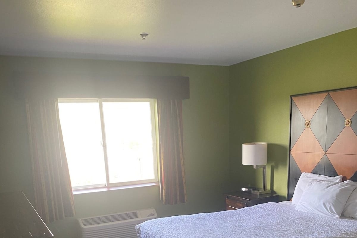 A hotel room with a large bed, a nightstand with a lamp, a window with curtains, and a ceiling light.
