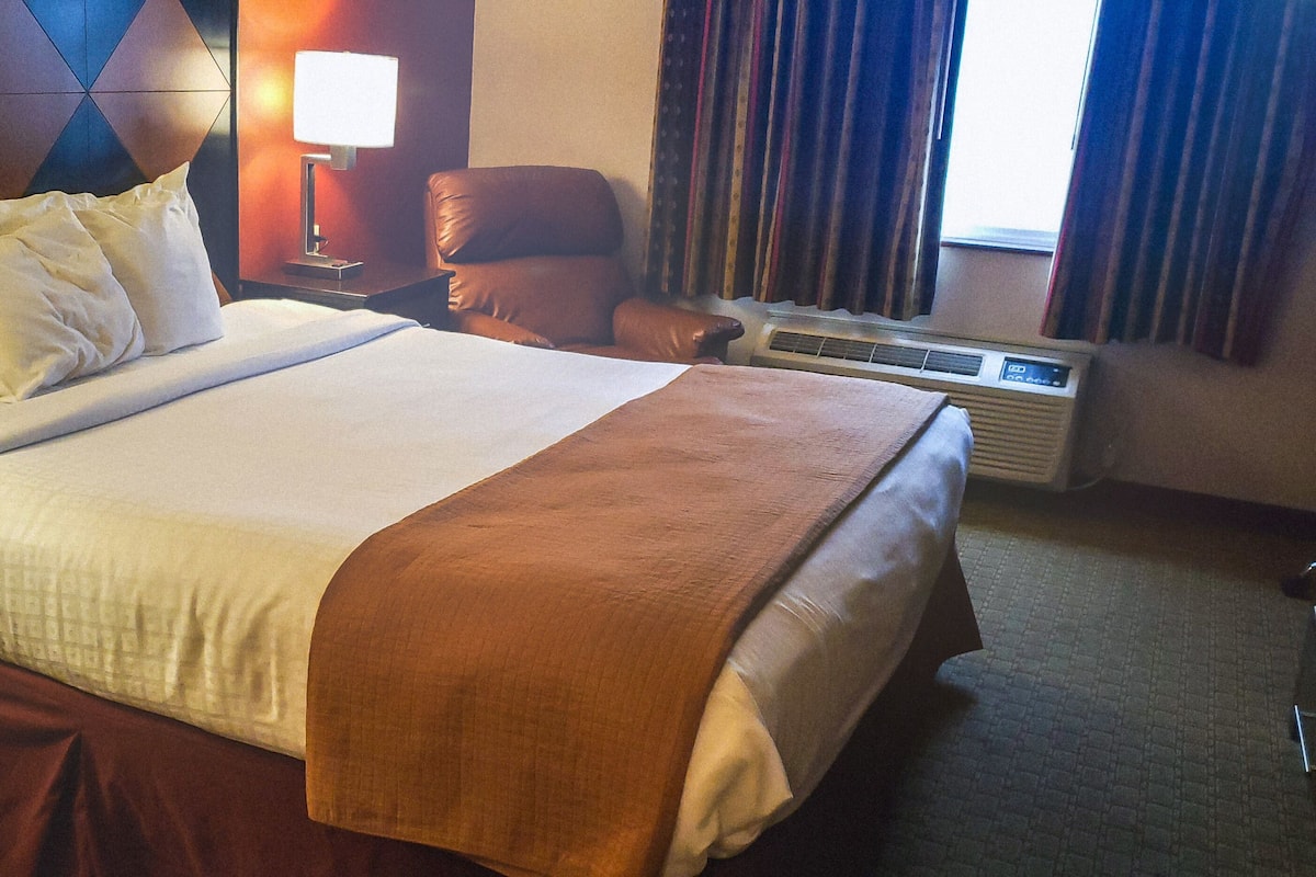 A hotel room with a large bed, a nightstand, a lamp, a chair, and an air conditioning unit.