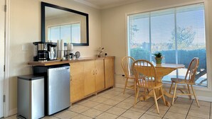 A compact kitchen with a coffee maker, cups, and a refrigerator. A dining table with chairs is positioned near a large window with blinds. - Snoozinn (Wilsonville)