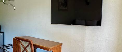 Quarto com escrivaninha e cadeira de madeira, televisão de tela plana na parede e uma pequena janela.
