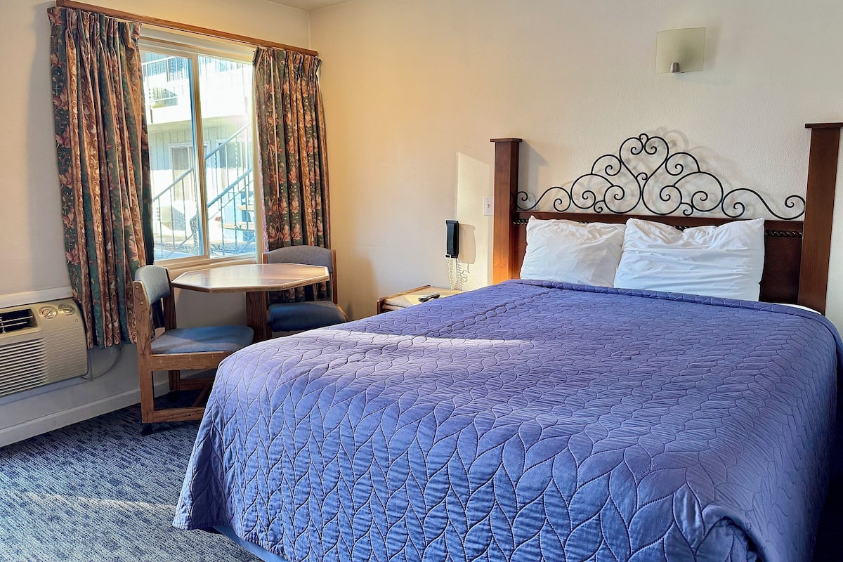 A hotel room with a large bed, a small table with a chair, a window with curtains, and a wall-mounted light fixture.