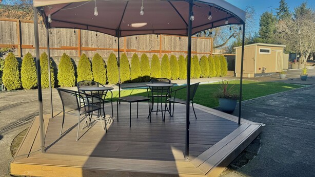 A covered patio area with a wooden deck, outdoor furniture, and a backdrop of neatly trimmed hedges. - Snoozinn (Wilsonville)