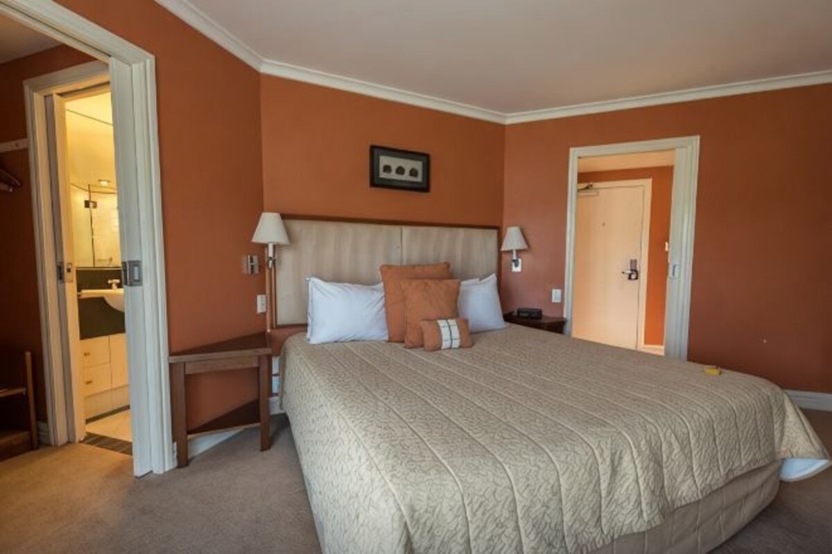 Habitación de hotel con una cama grande, mesitas de noche, una mesita auxiliar, una puerta y un cuadro enmarcado en la pared.