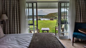 Une chambre avec un grand lit, une vue sur un espace verdoyant et un lac, visible à travers une porte vitrée, et une petite table avec des chaises à l’extérieur.