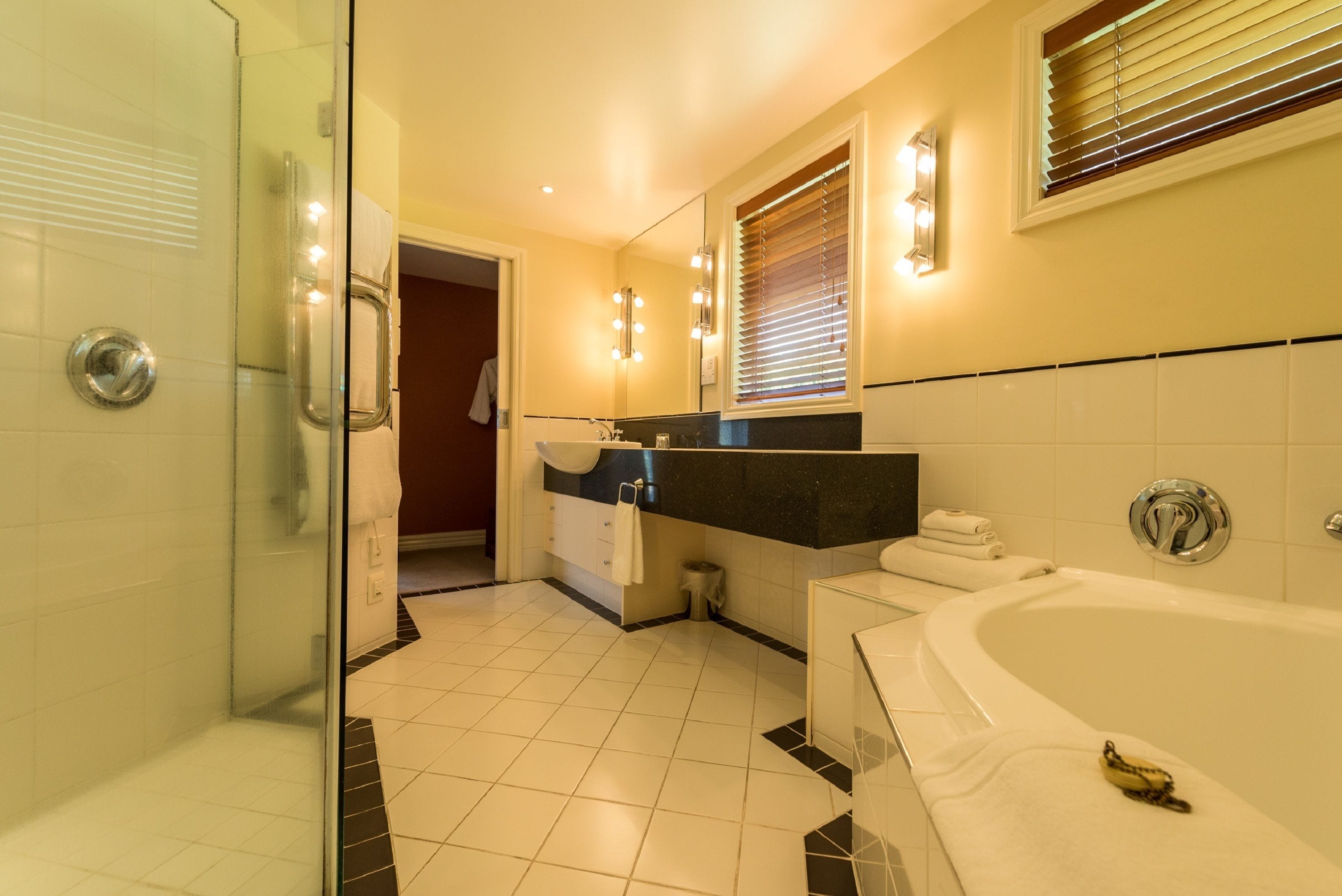 A modern bathroom with a glass shower enclosure, a bathtub, a vanity with a mirror, and a window with blinds.