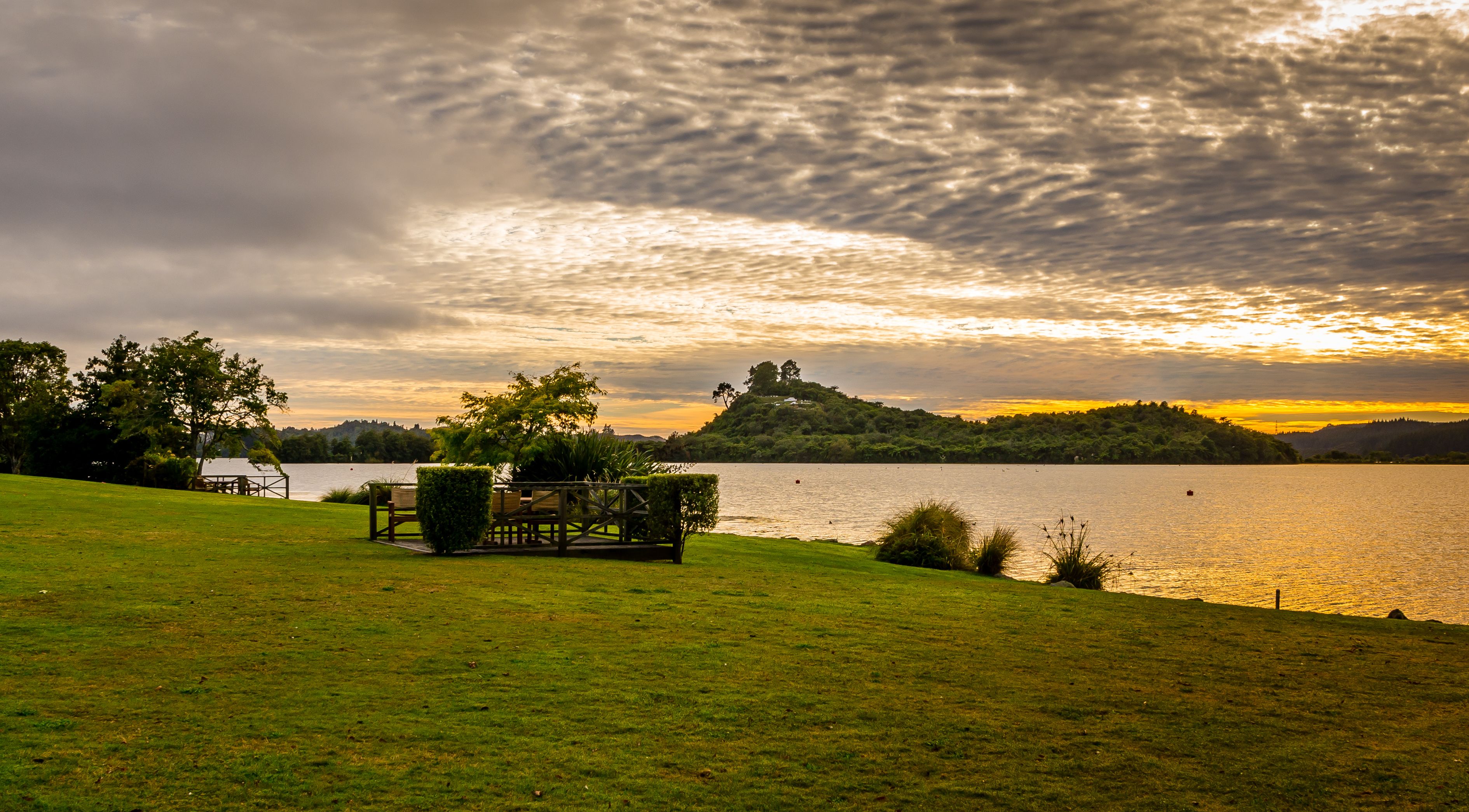 A grassy area with a wooden deck and benches overlooking a lake with a sunset in the background.