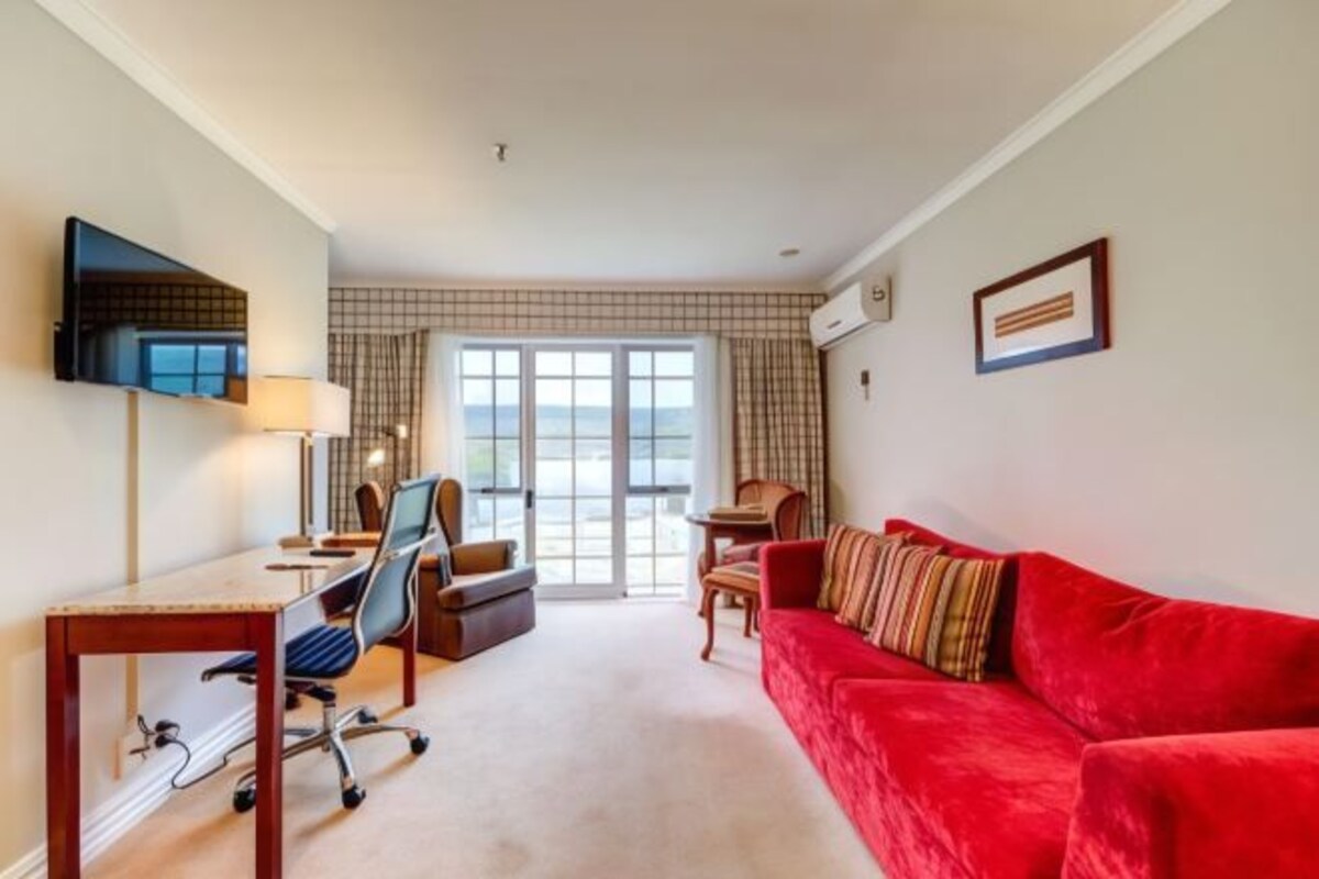 Habitación de hotel con sofá rojo, escritorio con silla, televisor montado en la pared y un ventanal con cortinas.