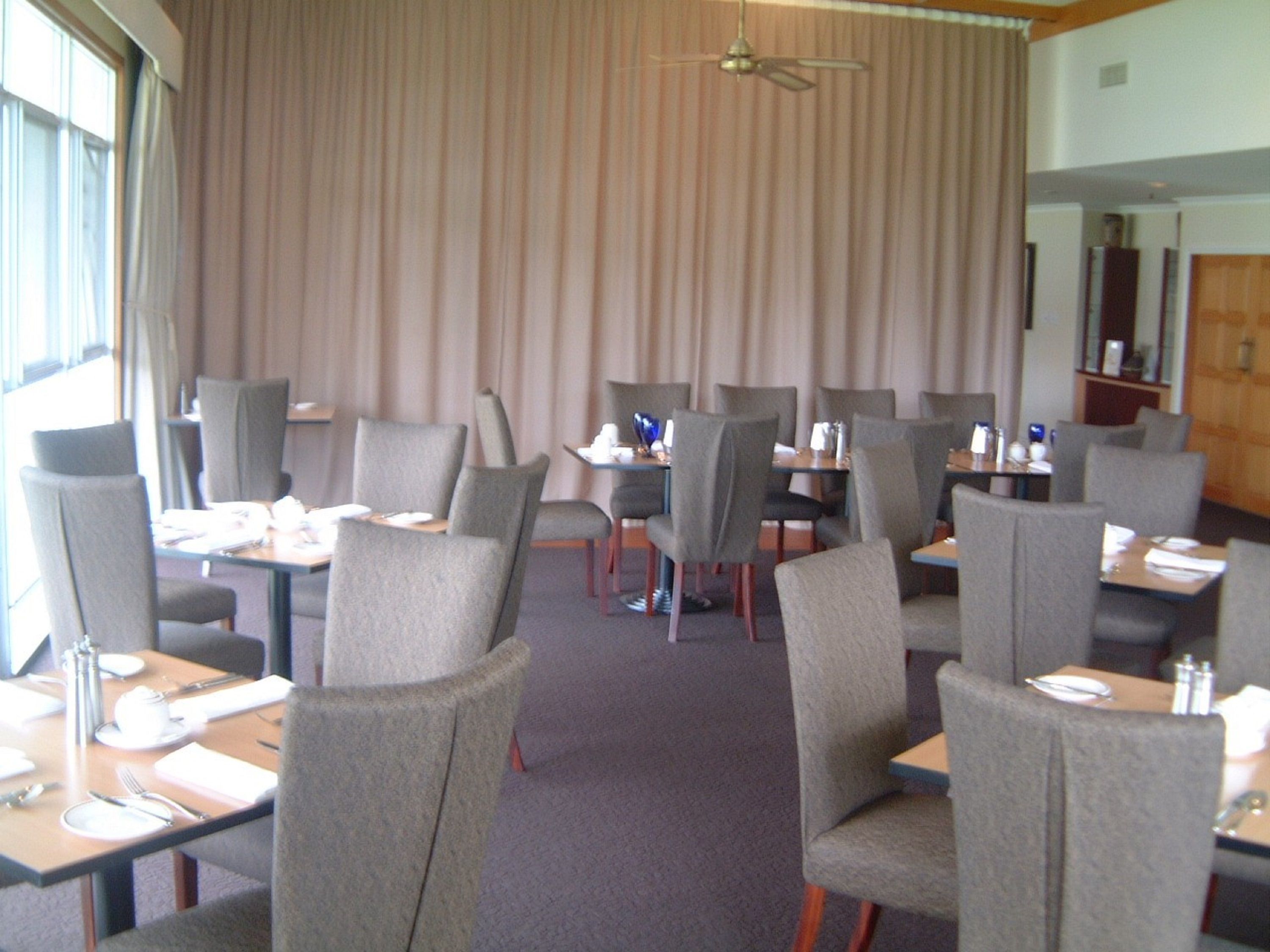 Une salle à manger d’hôtel avec plusieurs tables et chaises, un ventilateur de plafond et une grande fenêtre avec des rideaux.