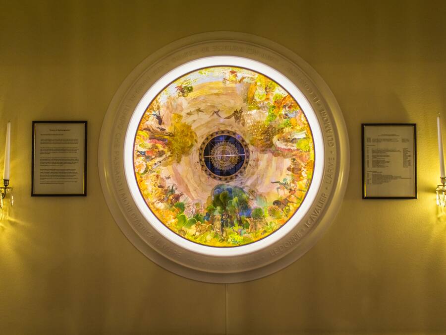 Ein kreisrundes Buntglasfenster mit einem zentralen Sonnendesign, das von zwei Wandleuchtern und zwei Infotafeln gesäumt wird.