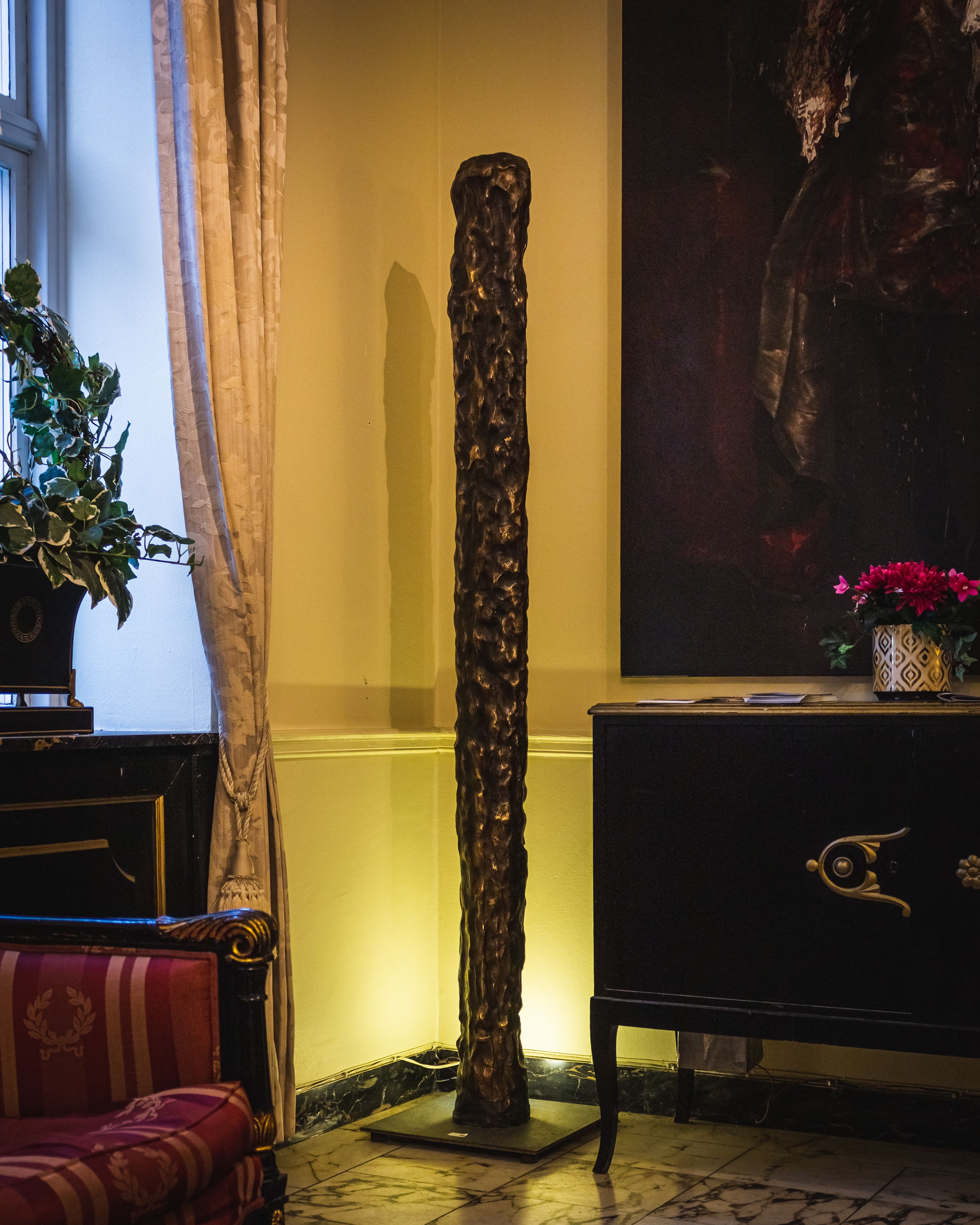 A room with a tall, textured sculpture, a painting on the wall, a potted plant, and a patterned sofa.