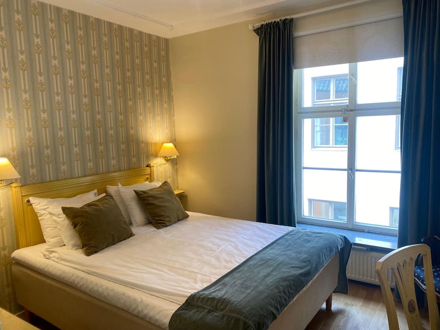 A hotel room with a bed, two bedside lamps, a window with curtains, and a radiator.