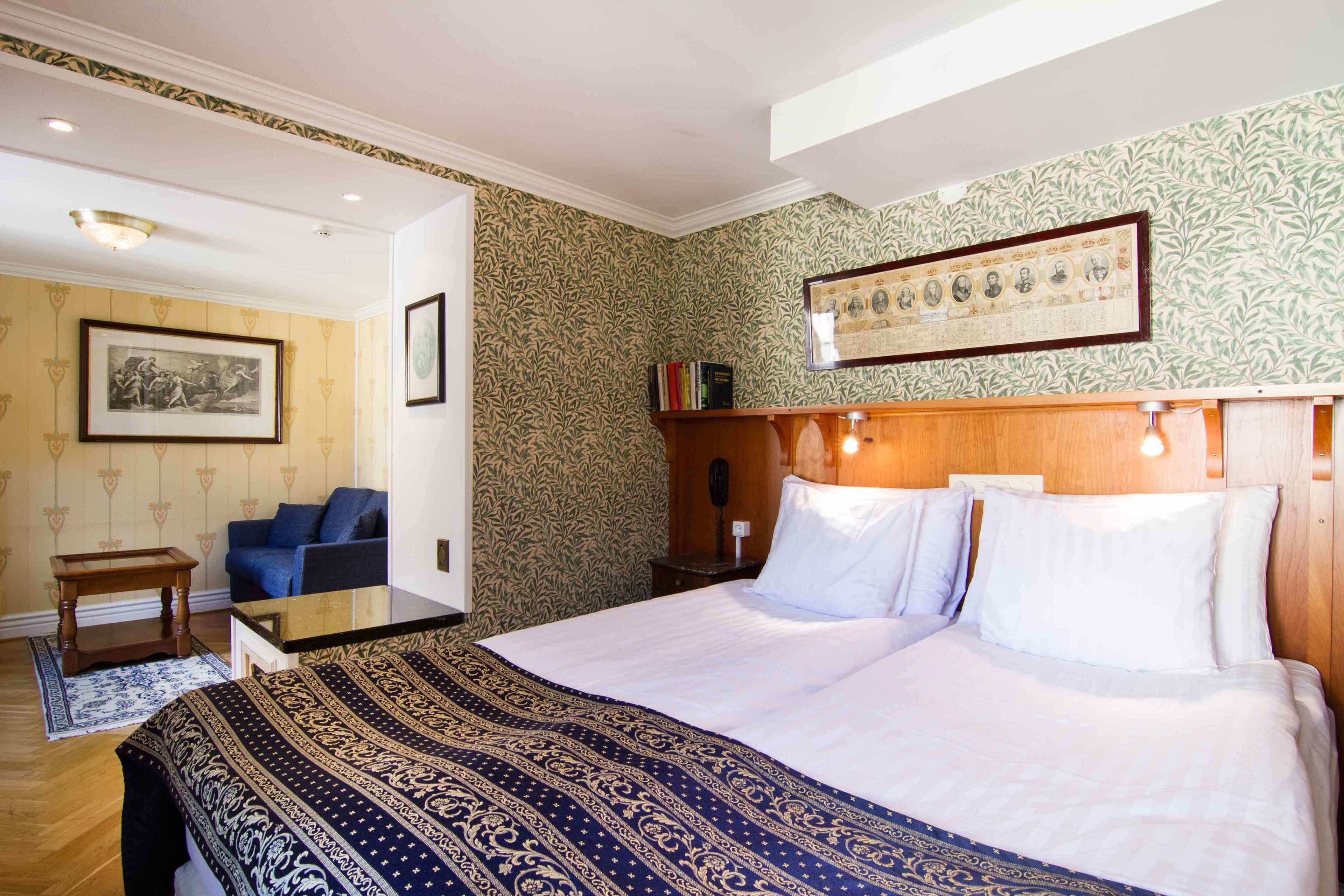 A hotel room with a large bed, a wooden headboard, a small table, a sofa, and a patterned wall.