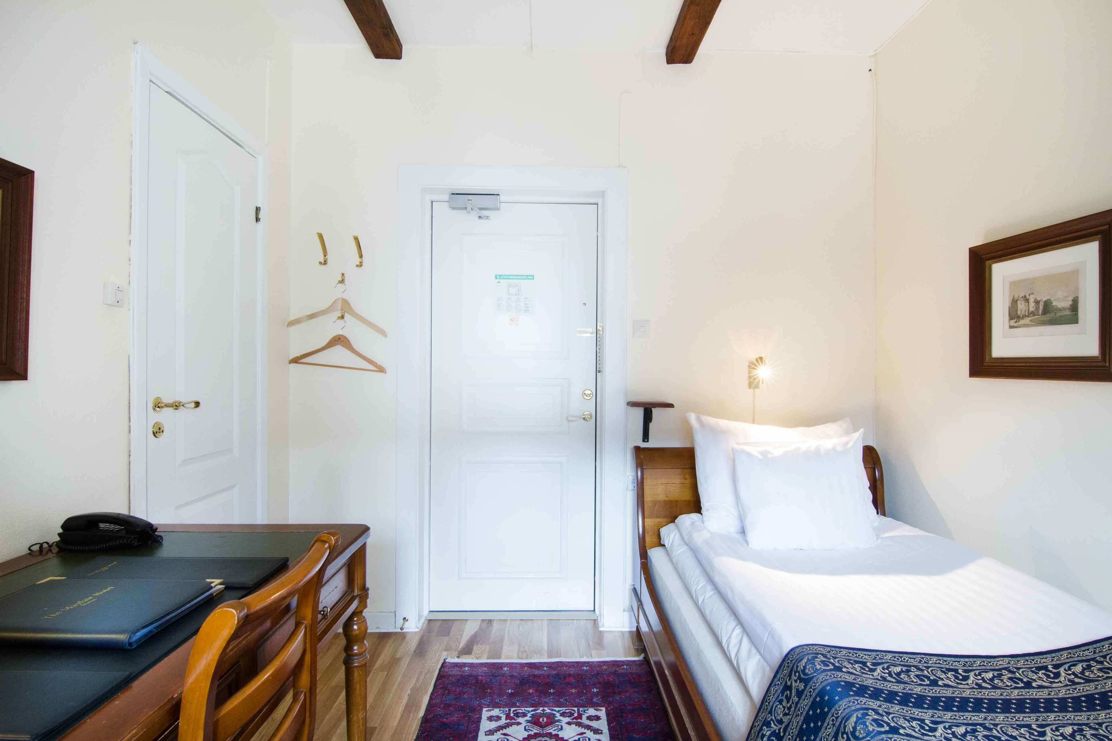 A hotel room with a bed, a desk with a chair, a door, and a small table with a phone and a folder.