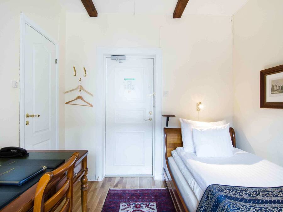 A hotel room with a bed, a desk with a chair, a door, and a small table with a phone and a folder.