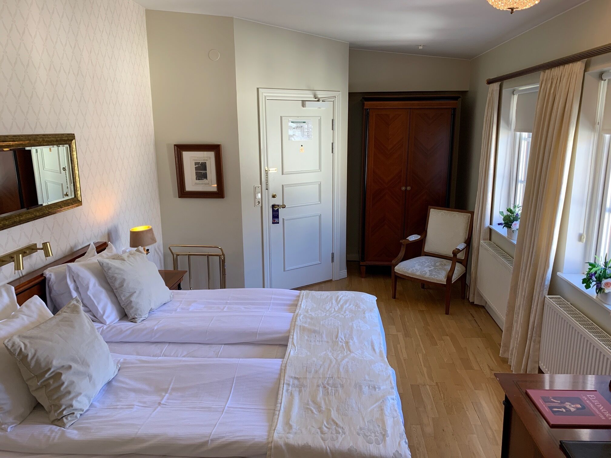 A hotel room with two beds, a chair, a desk, a radiator, a door, and a window with curtains.