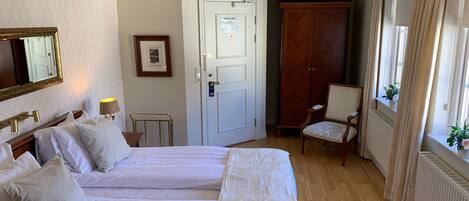 A hotel room with two beds, a chair, a desk, a radiator, a door, and a window with curtains.