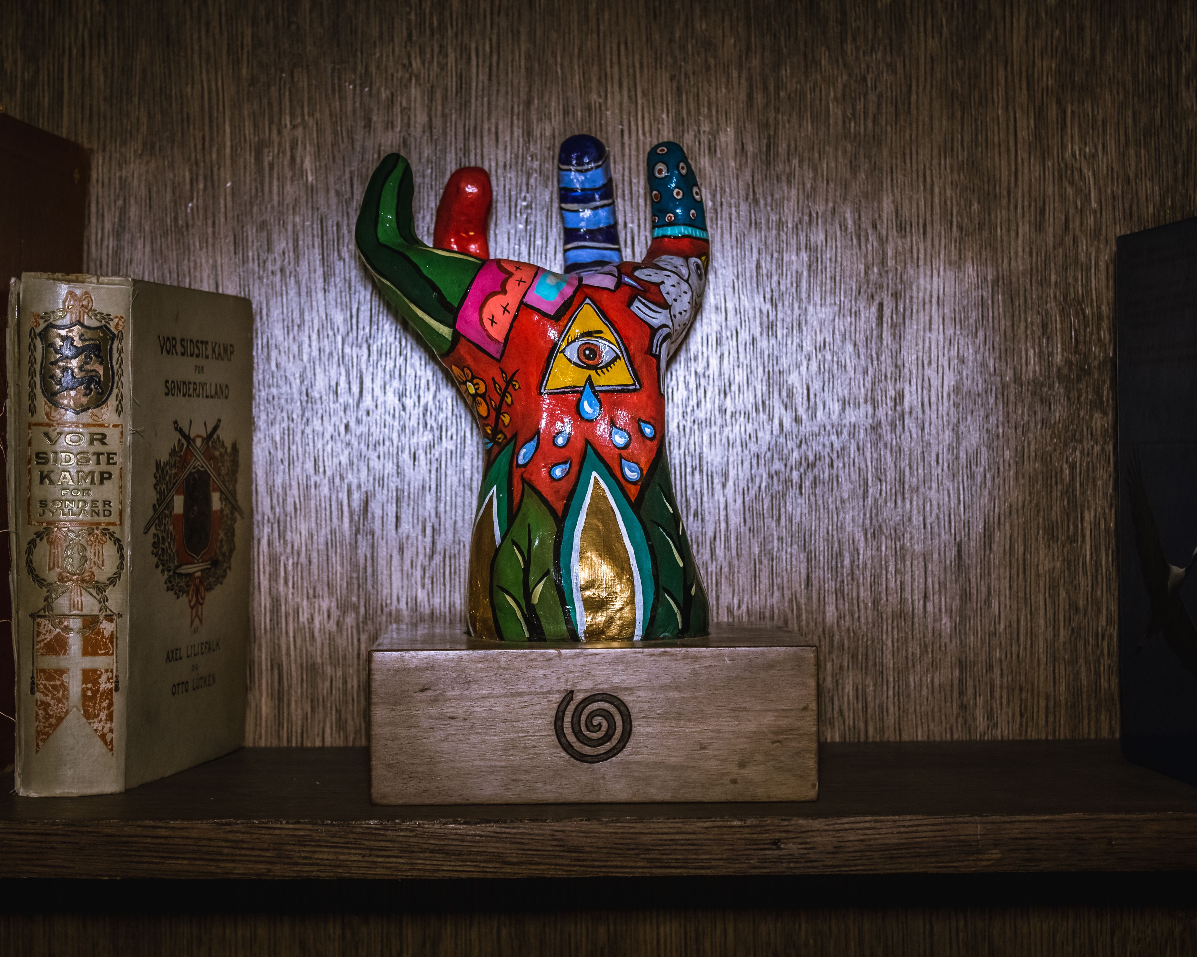 En hylla i trä med en färgstark skulptur formad som en hand och en bok med titeln "Vor Sidsste Kamp for Sondersyldland" av Axel Liljerup.
