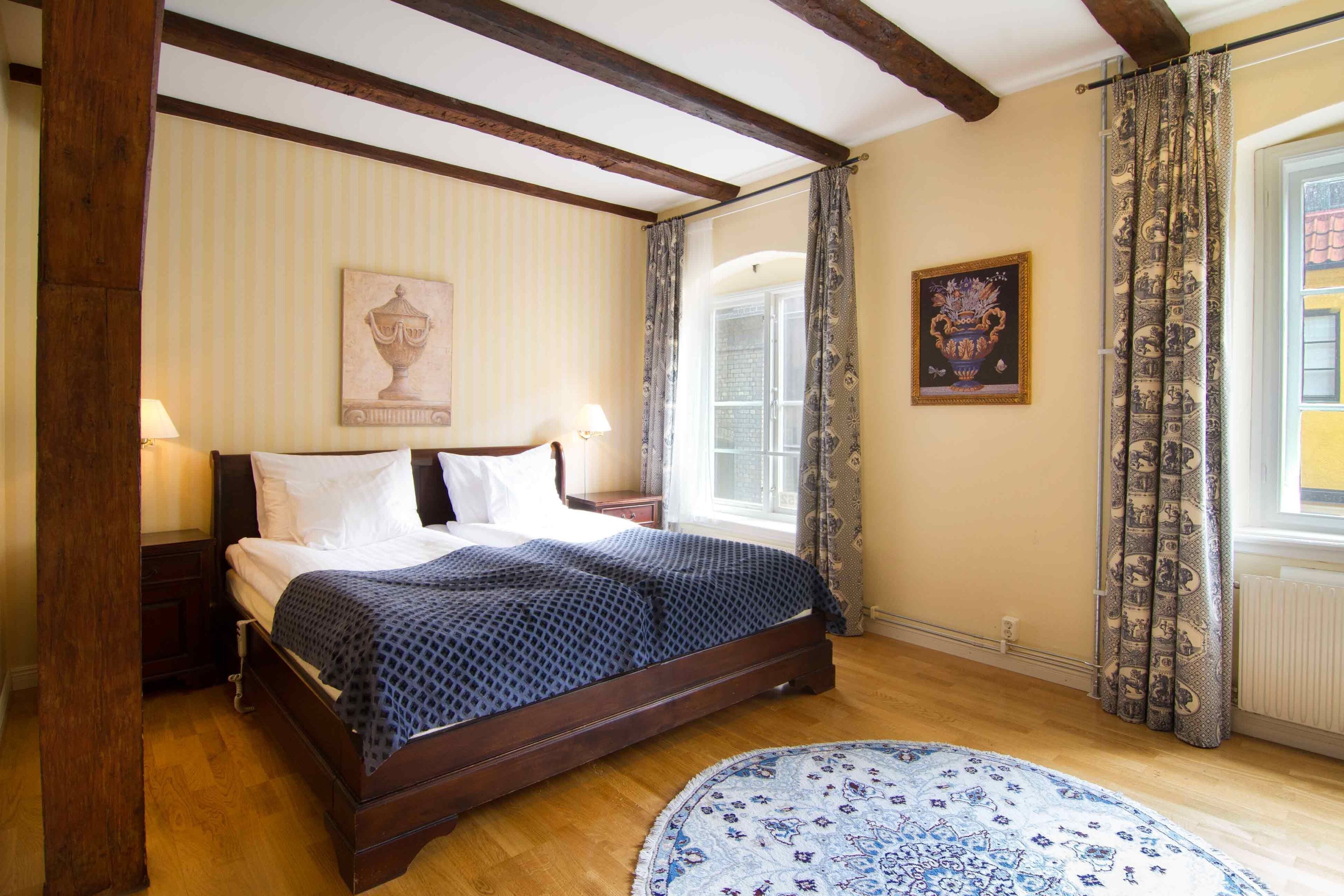 Un dormitorio con una cama de madera, un tapete redondo, una ventana con cortinas e imagen colgada en la pared.