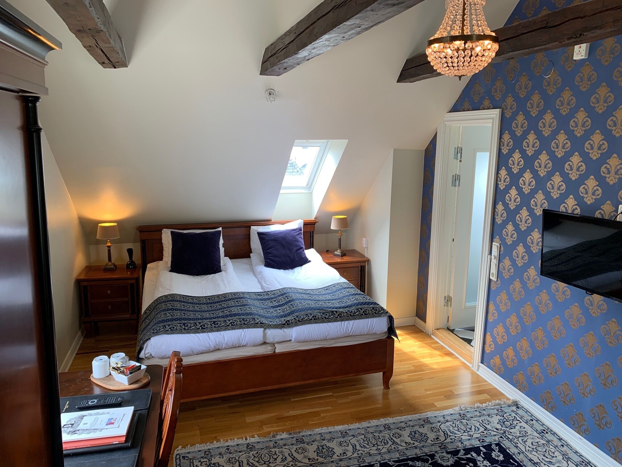 A bedroom with a large bed, wooden headboard, two bedside tables with lamps, a TV mounted on the wall, and a patterned carpet.