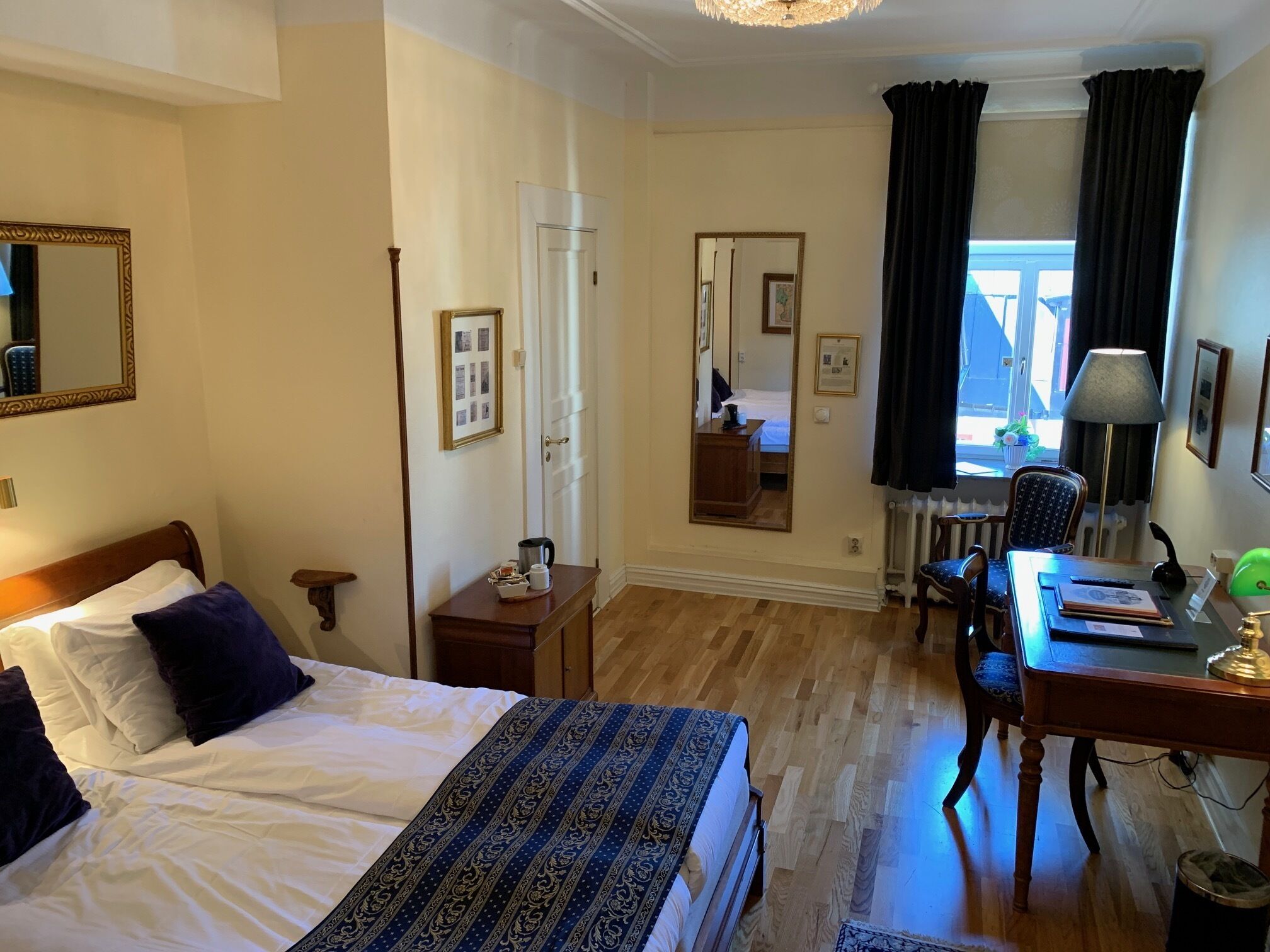Hotelkamer met een bed, een bureau, een stoel, een lamp, een spiegel en een raam met gordijnen.