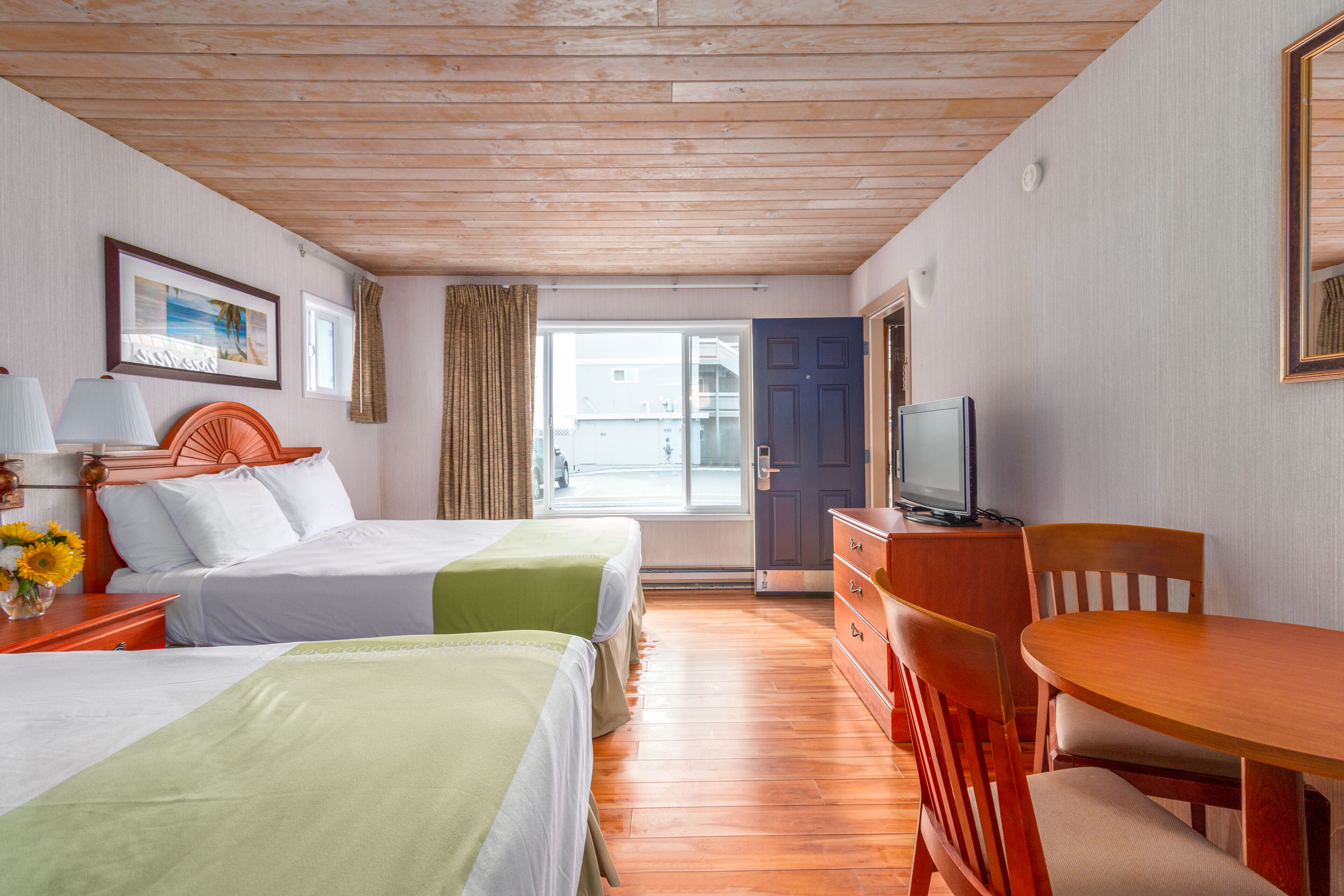 A hotel room with two beds, a wooden ceiling, a TV, a dining table, and a view through a glass door.