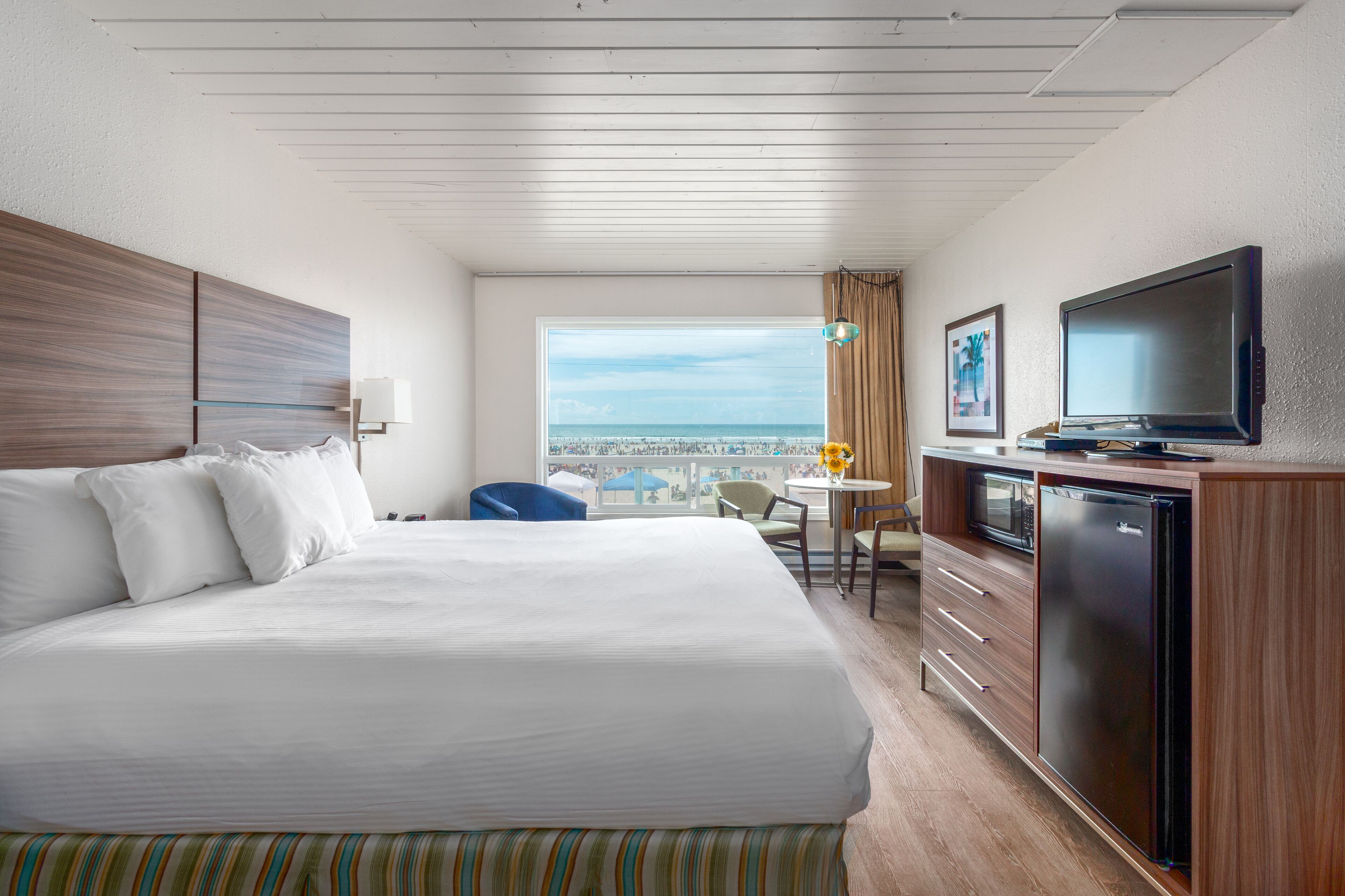 Habitación de hotel con una cama grande, un escritorio con televisor, una mesita con lámpara y vista a la playa.