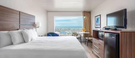 Habitación de hotel con una cama grande, un escritorio con televisor, una mesita con lámpara y vista a la playa.