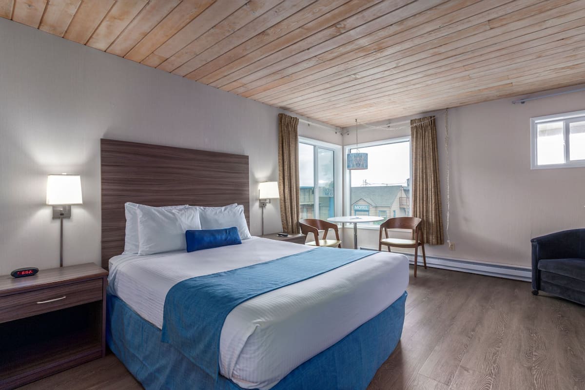 Habitación de hotel con techo de madera, una cama grande con ropa de cama blanca y azul, dos lámparas de noche, una pequeña mesa con sillas y vistas al exterior a través de una ventana.