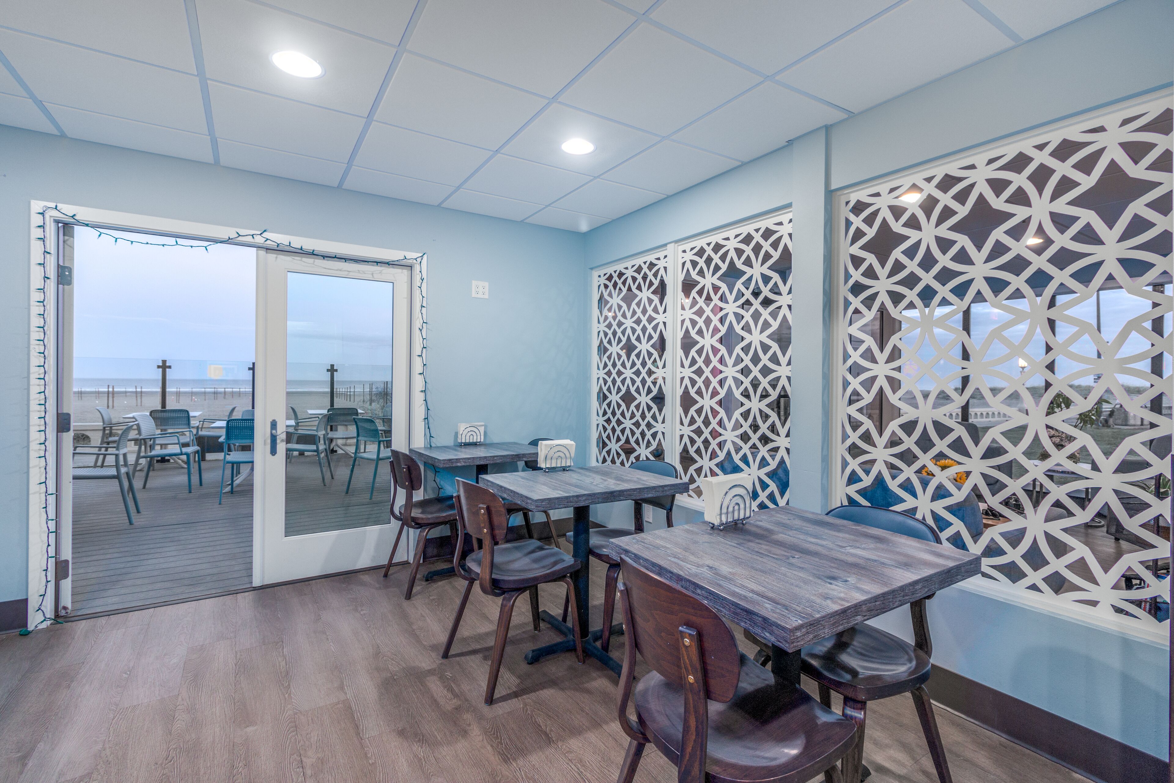 Un comedor con mesas y sillas, una puerta de vidrio que da al exterior, y paneles de celosía blanca decorativos.