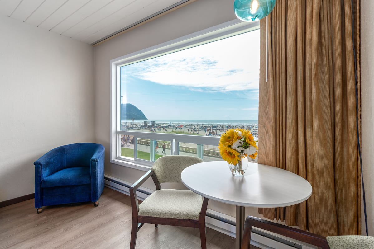 Un balcón con vista al mar, una silla azul, una mesa redonda con un jarrón de flores y una cortina.