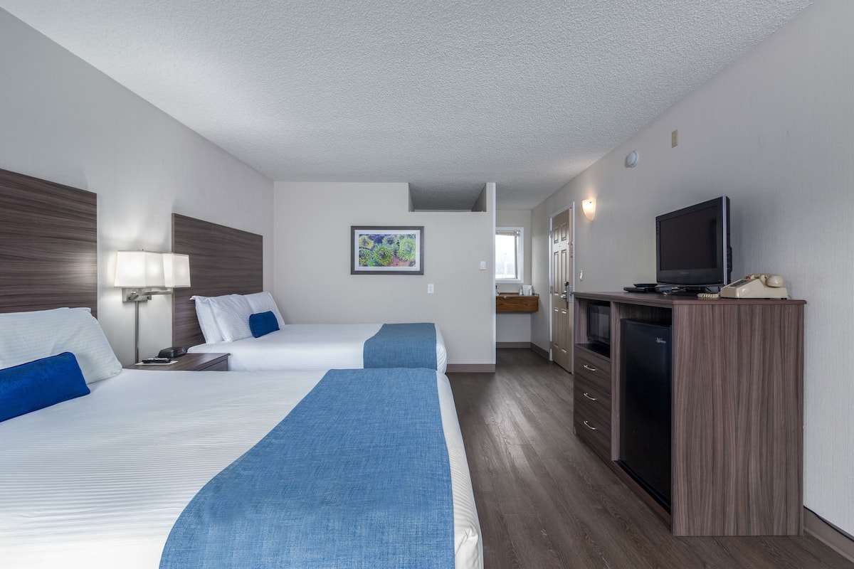 Habitación de hotel con dos camas, televisión, escritorio con computadora y una kitchenette pequeña.