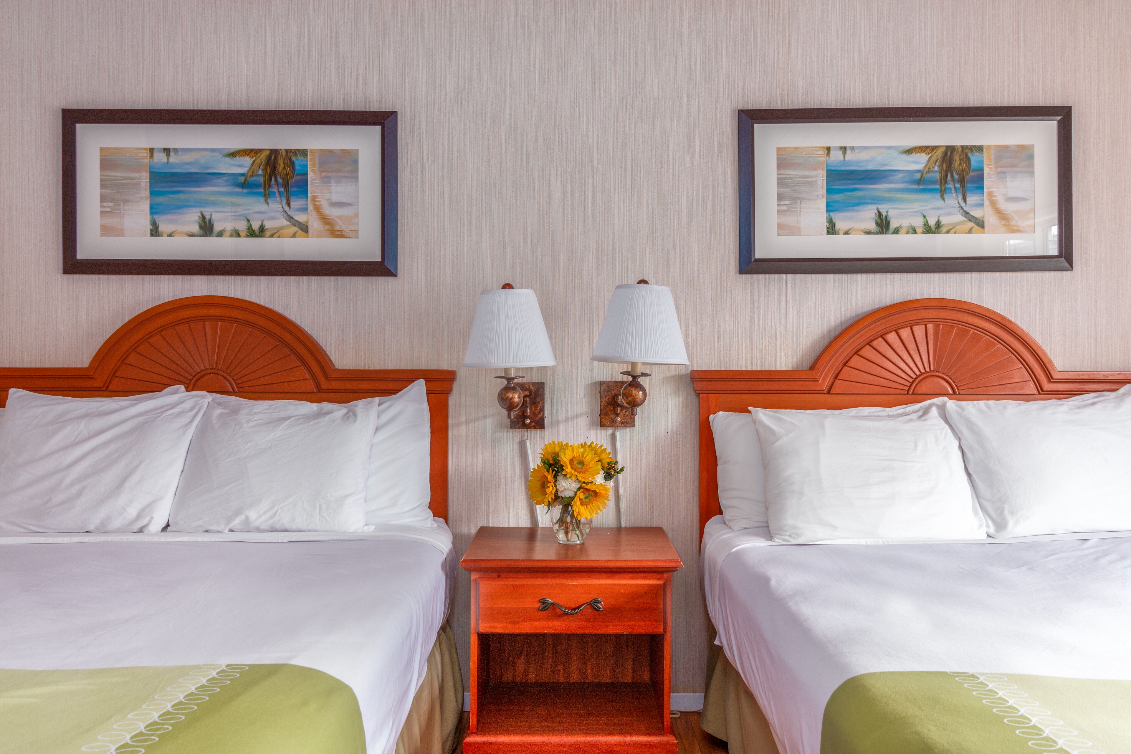 A hotel room with two beds, a nightstand with a vase of flowers, and framed artwork on the wall.