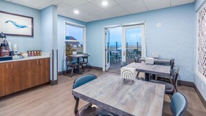 A modern dining area with wooden tables and chairs, a kitchen counter with various items, and large windows offering a view of the city. - Seashore Inn on the Beach (Seaside)