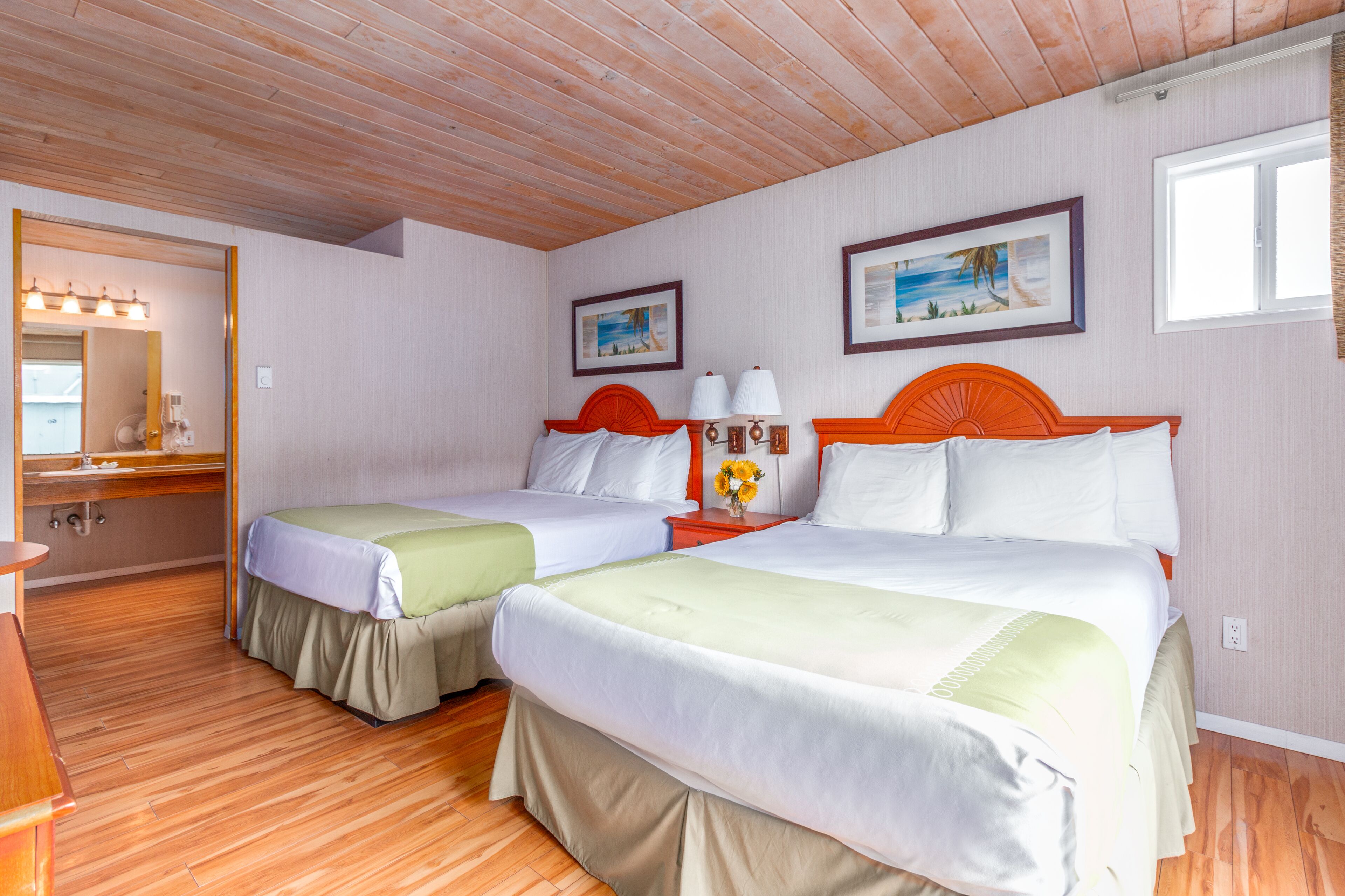 A hotel room with two beds, wooden flooring, a wooden ceiling, a bathroom with a mirror, and framed pictures on the wall.