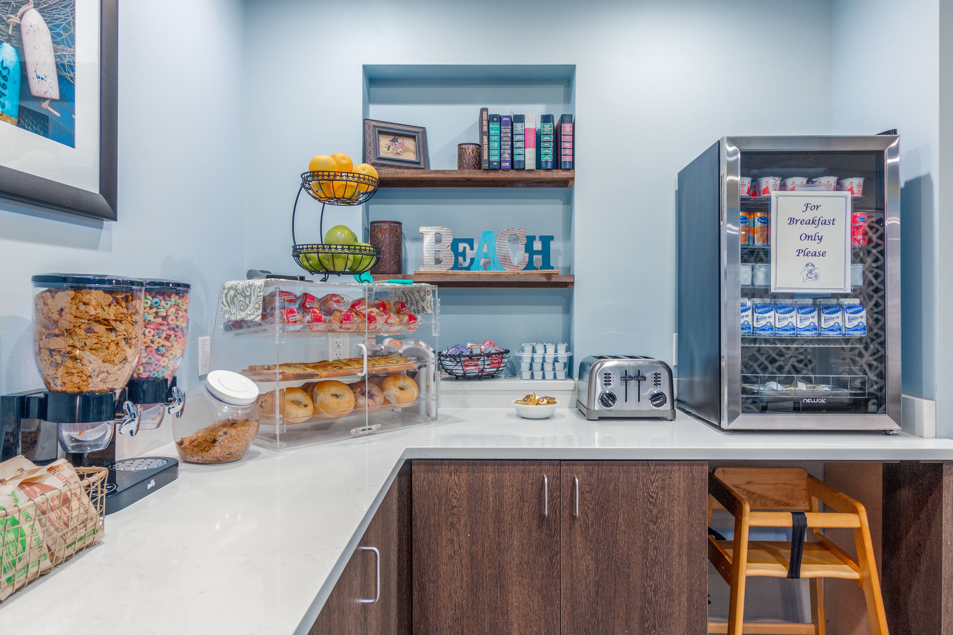 A breakfast bar with a coffee machine, a toaster, and a beverage dispenser. There are shelves with books and a sign that says "For Breakfast Only Please".