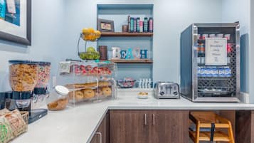 A breakfast bar with a coffee machine, a toaster, and a beverage dispenser. There are shelves with books and a sign that says "For Breakfast Only Please".