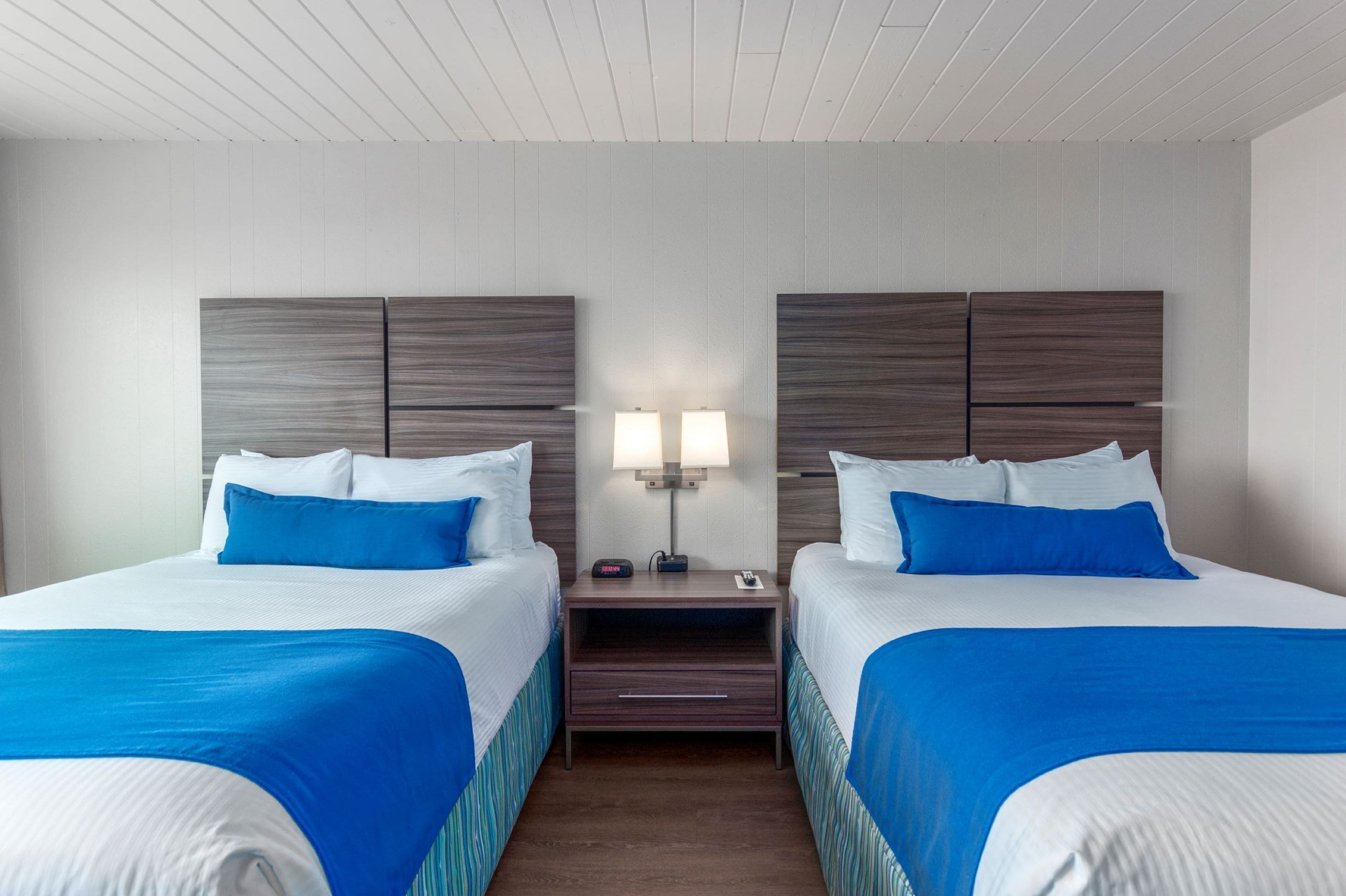 A hotel room with two beds, each with a blue bedspread and white pillows. There is a nightstand with a lamp between the beds.