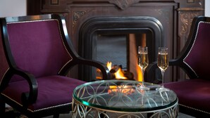 A room with two purple upholstered chairs, a glass table with two champagne glasses, and a fireplace in the background.