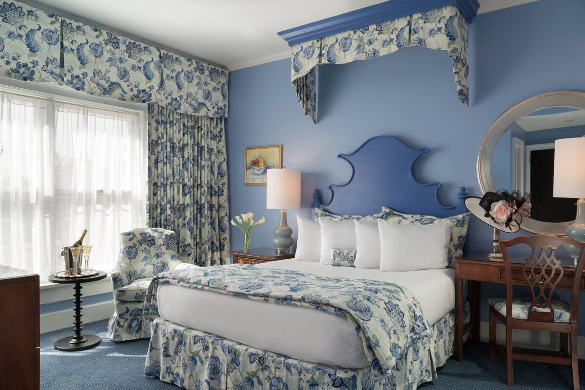 Un dormitorio con una colcha estampada con motivos florales, un espejo grande y una silla con cojín floral.