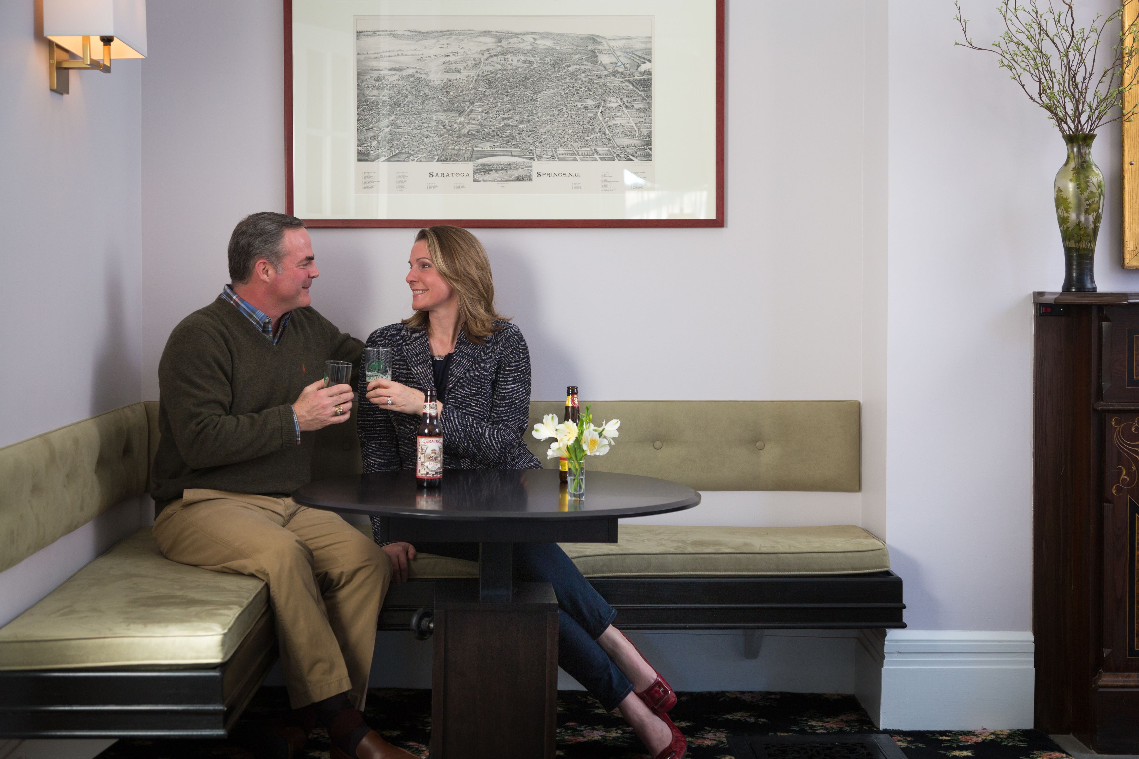 A man and a woman sitting on a bench, holding glasses, with a framed map on the wall behind them.
