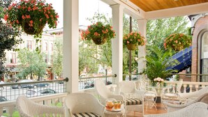 A covered outdoor seating area with wicker furniture, hanging flower baskets, and a view of the street.